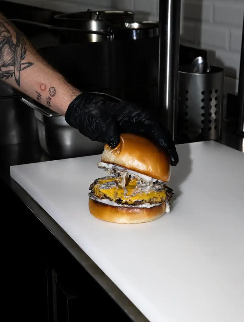 Mutfakta eldivenli bir el, çift katlı smash burger’ın üst ekmeğini yerleştiriyor.